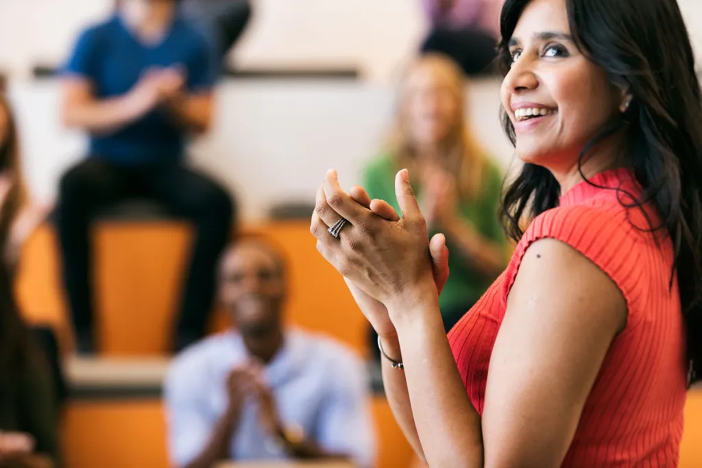 A woman in a red dress clapping and smiling in an auditorium, with a blurred background of people applauding.