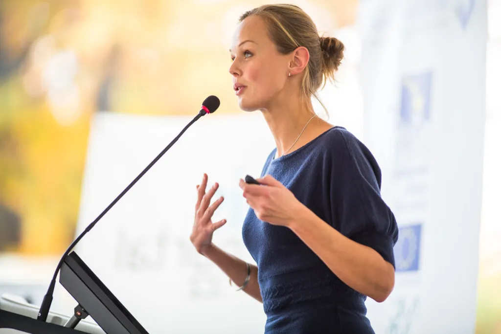 A woman in a navy dress speaking passionately at a podium with a microphone, gesturing with her hands while delivering a presentation.