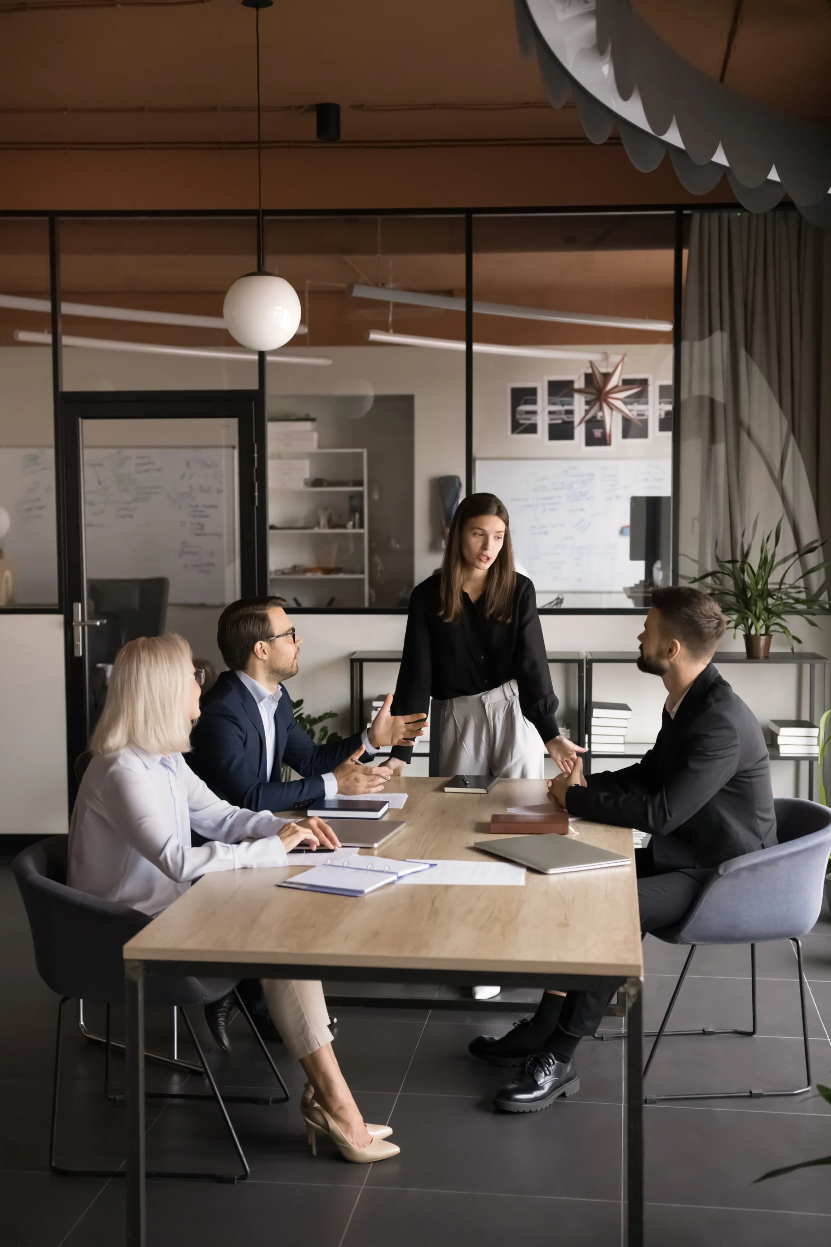 A professional meeting in a modern office, with a young woman standing and presenting to a group of seated colleagues engaged in discussion.