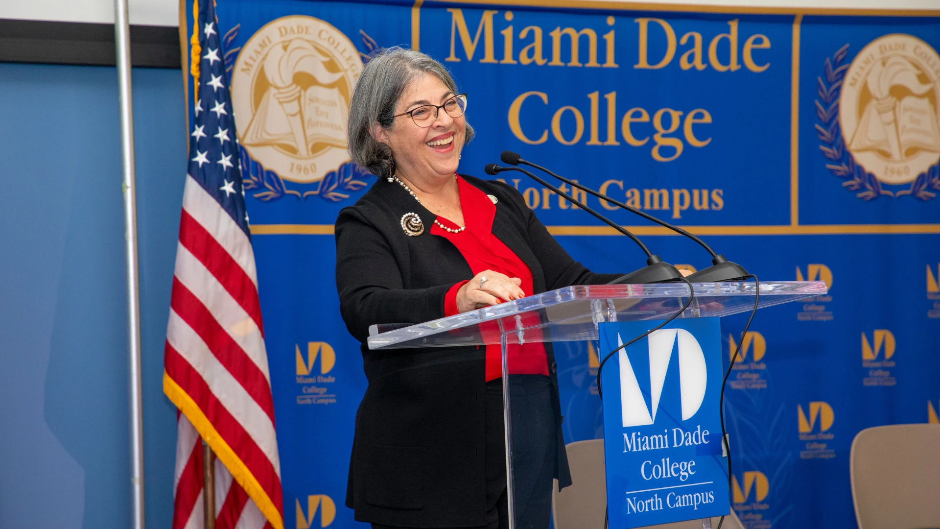 A woman speaking at a podium at Miami Dade College North Campus, smiling while addressing an audience, with an American flag in the background.