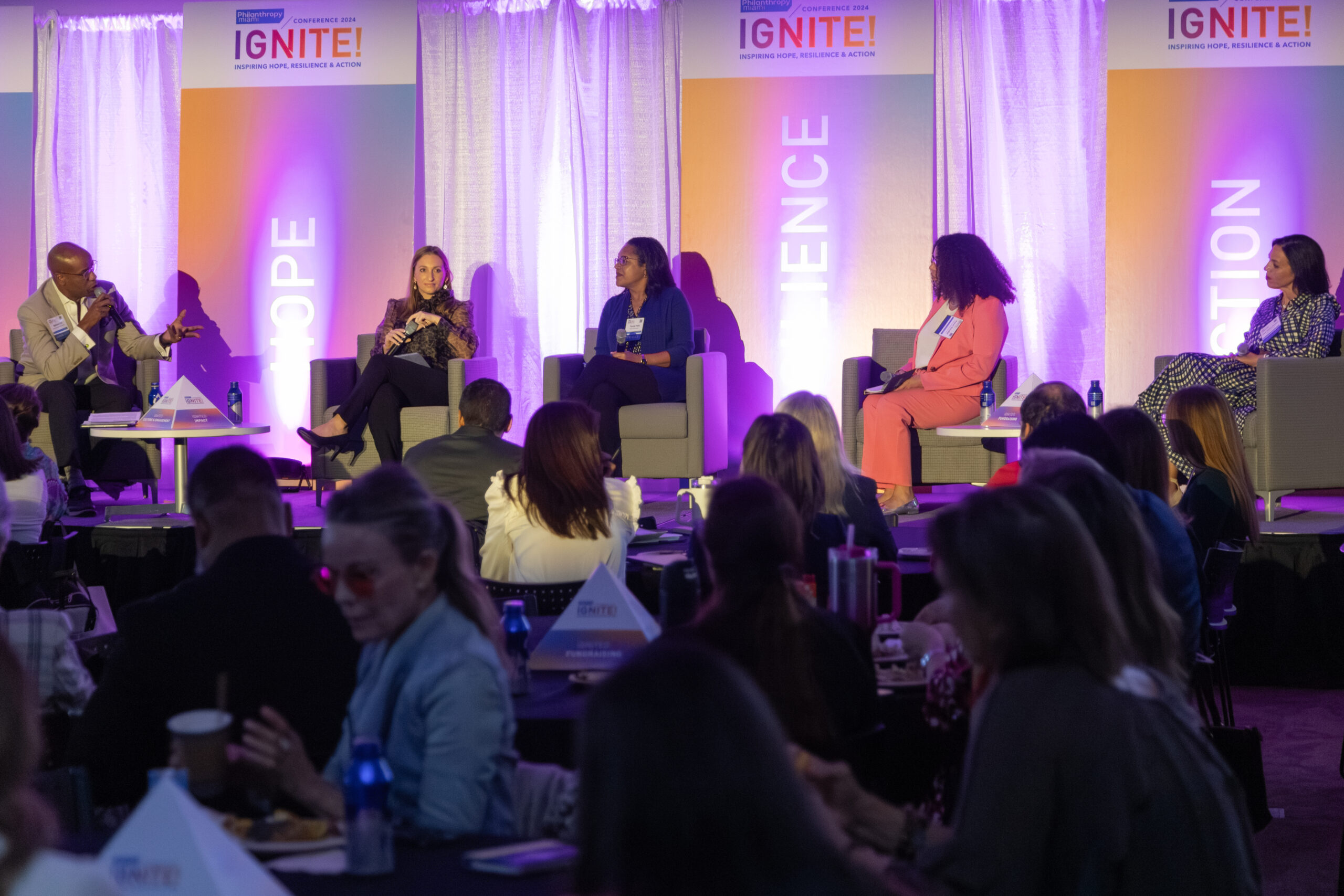 A panel discussion at the Ignite! Conference with five speakers seated on stage, engaging in conversation, while an audience listens attentively.