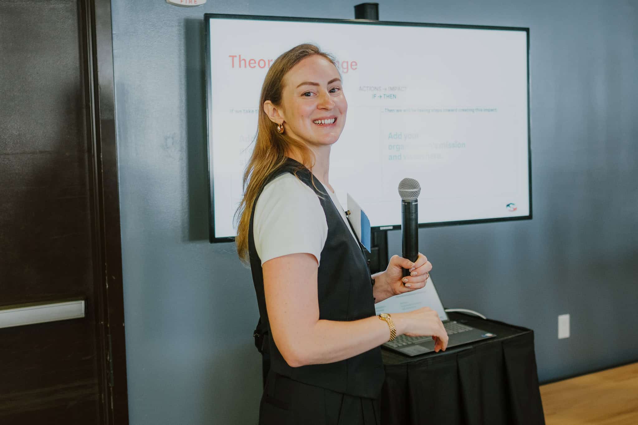 A woman giving a presentation, holding a microphone and smiling, standing in front of a screen displaying a slide with text.