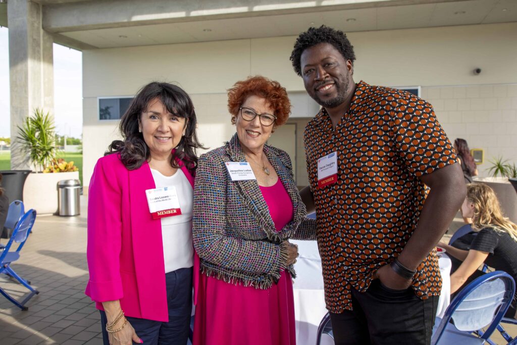 Three people smiling at an outdoor networking event, wearing name tags and business attire, with a casual and welcoming atmosphere.