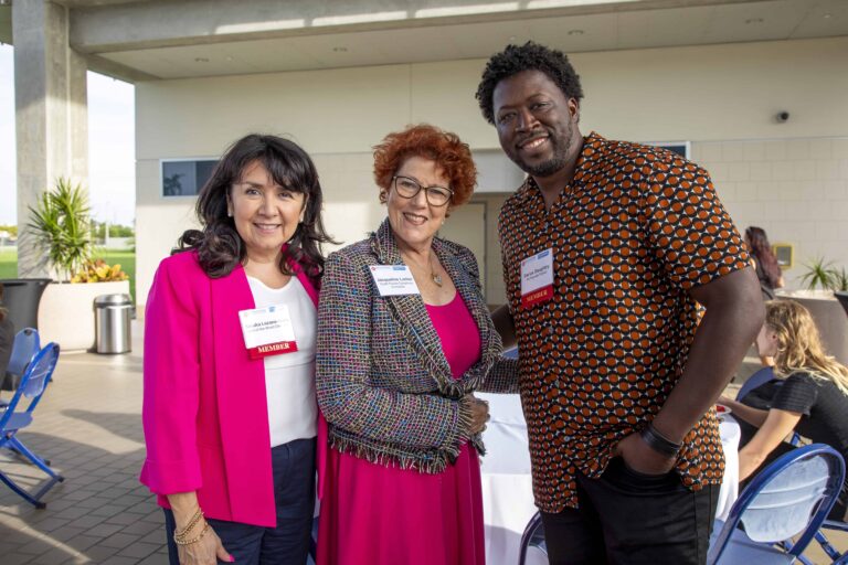Three people smiling at an outdoor networking event, wearing name tags and business attire, with a casual and welcoming atmosphere.