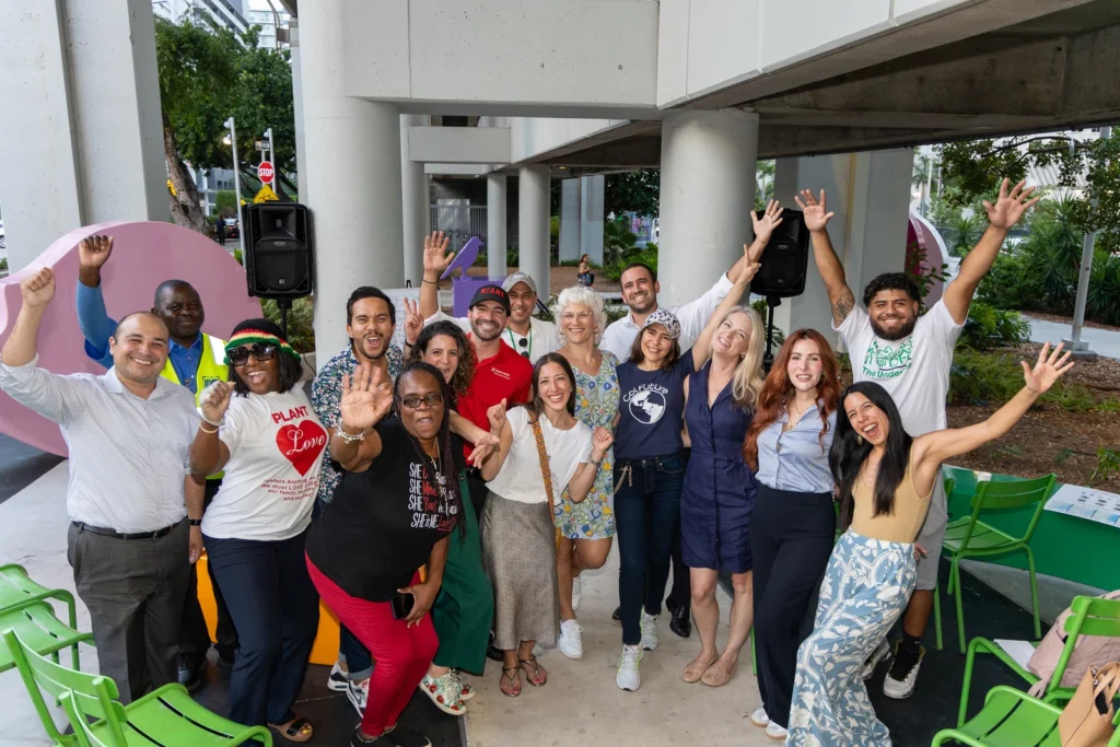 A diverse group of people outdoors posing with excitement, raising their hands and smiling in a vibrant, energetic setting.