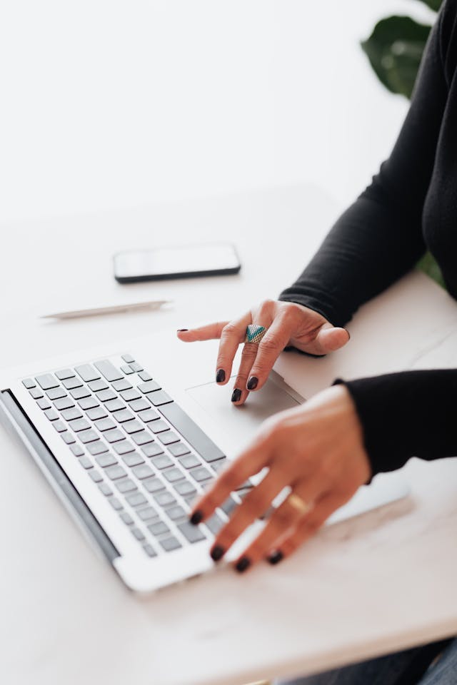 A close-up of a person's hands with dark nail polish typing on a laptop, with a smartphone and a pen on the desk.