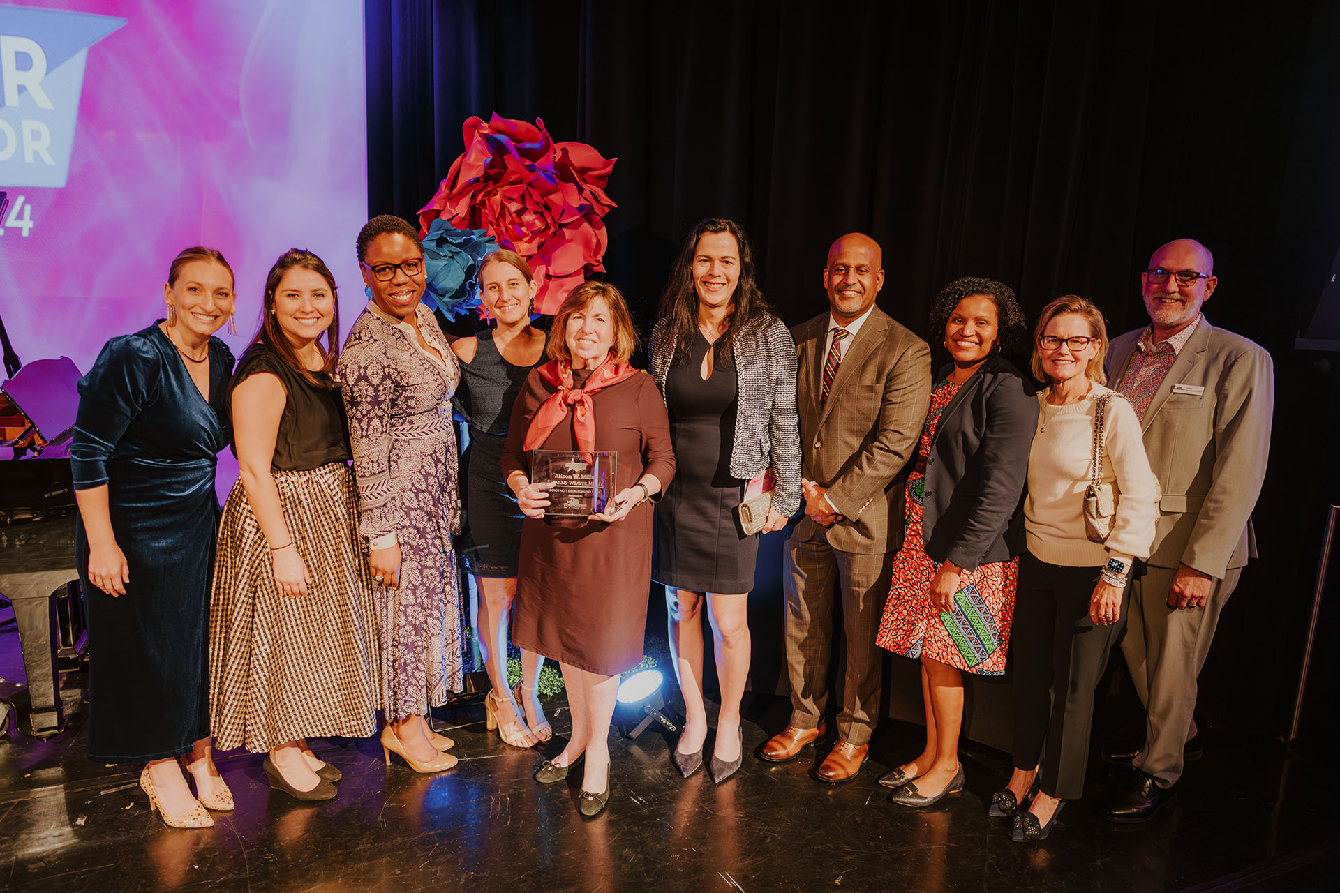 A diverse group of professionally dressed individuals stands on a stage, smiling, with a woman in the center holding an award.